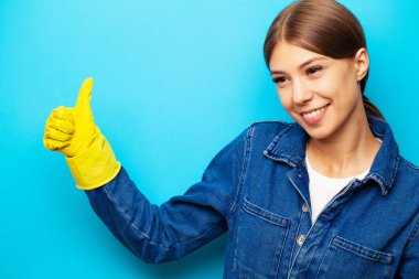 Önlüklü ve sarı eldivenli bir temizlik şirketinin çalışanı mavi arka planda poz veriyor.