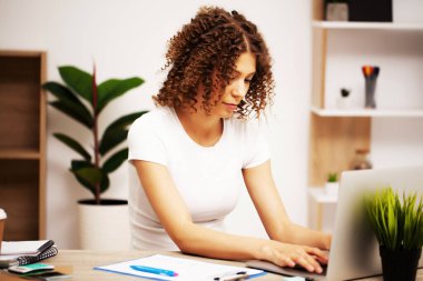 Kafasında bukleler olan yorgun bir kadın ofiste dizüstü bilgisayarla çalışıyor.