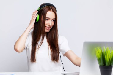 Kulaklıkla müzik dinleyen genç ve güzel bir kadın ofiste dizüstü bilgisayarla çalışıyor.