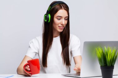 Kulaklıkla müzik dinleyen genç ve güzel bir kadın ofiste dizüstü bilgisayarla çalışıyor.