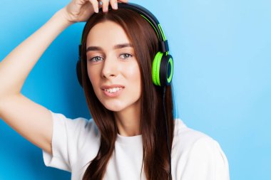 Güzel genç kadın model mavi arka planda kulaklıkla müzik dinliyor.