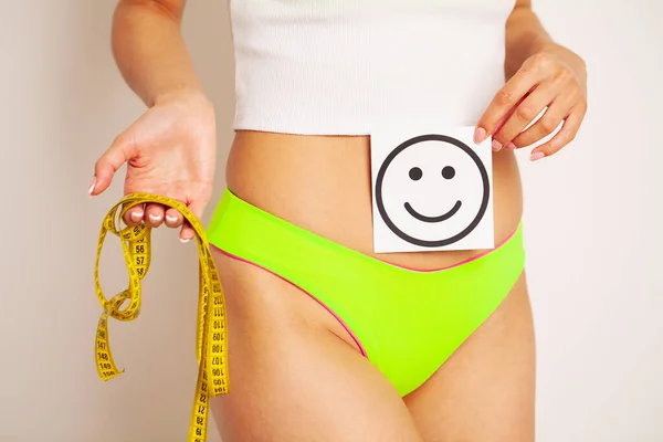 Primer plano de una mujer con una figura delgada demuestra el resultado sosteniendo una tarjeta cerca de su estómago con una sonrisa sonriente y cinta métrica amarilla. — Foto de Stock