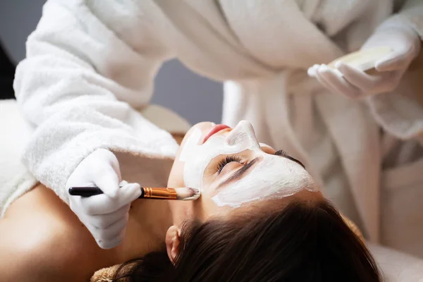 Estetista applica una maschera sul viso di una donna sdraiata su un divano in una spa — Foto Stock