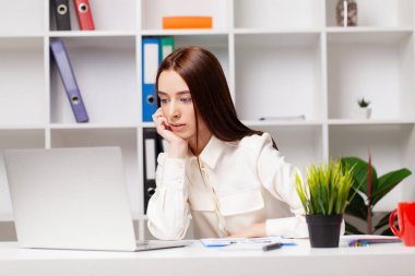 Genç ve güzel bir iş kadını, modern ofiste dizüstü bilgisayarla çalışıyor.