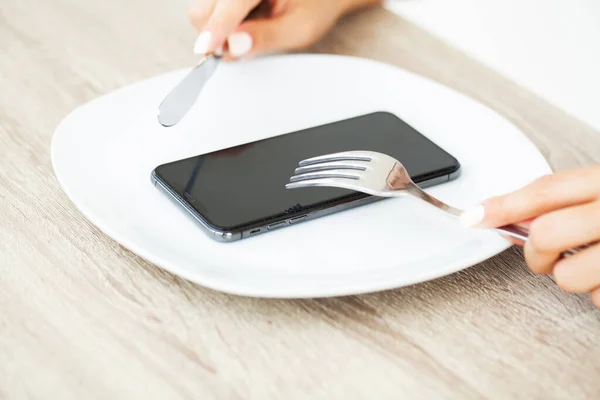 Dependence on the phone, close up a womans hands holding a fork and a knife over the phone on plate — Stock Photo, Image