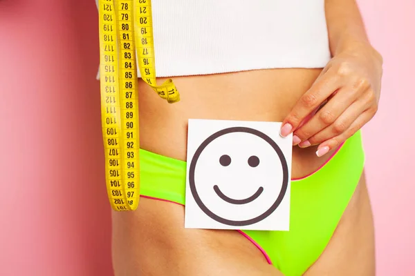 Primer plano de una mujer con una figura delgada demuestra el resultado sosteniendo una tarjeta cerca de su estómago con una sonrisa sonriente y cinta métrica amarilla — Foto de Stock