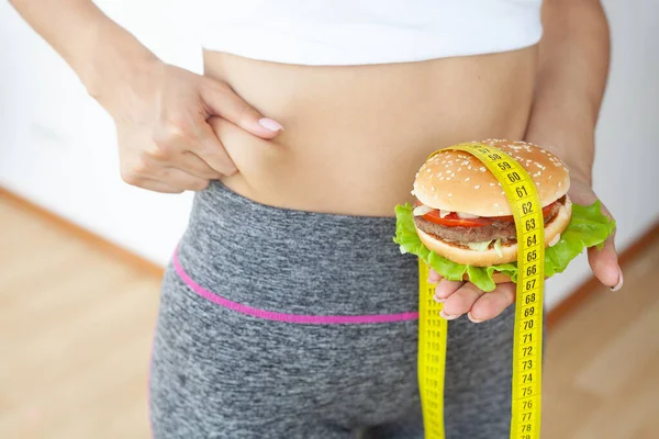 Concepto de dieta, mujer sosteniendo hamburguesa grasa dañina con cinta métrica amarilla —  Fotos de Stock