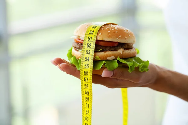 Ernährungskonzept: Frau hält schädlichen Fett-Burger mit gelbem Maßband — Stockfoto