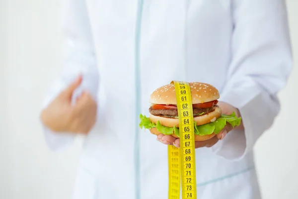 Диетическая концепция, доктор держит вредный жирный бургер с желтой измерительной лентой — стоковое фото