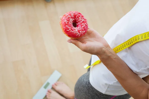 Ernährungskonzept: Frau misst Gewicht auf elektronischer Waage und hält Kalorienkrapfen in der Hand — Stockfoto