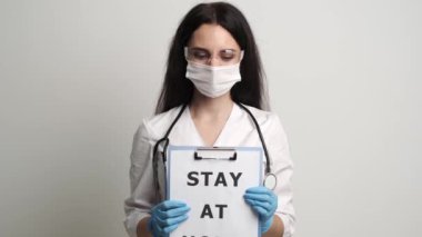 Maskeli ve eldivenli doktor, üzerinde yazı olan bir form tutuyor. Evde kalın.