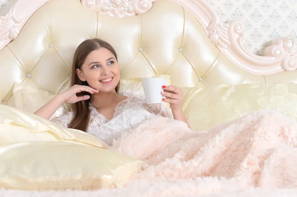 コーヒーを飲みながら自宅のベッドで横になっている若い女性 — ストック写真