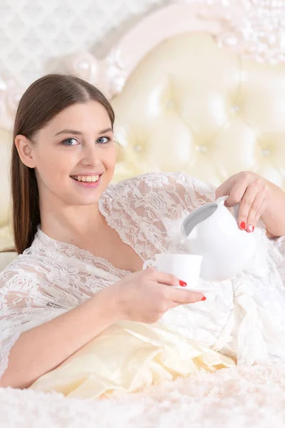 コーヒーを飲みながら自宅のベッドで横になっている若い女性 — ストック写真