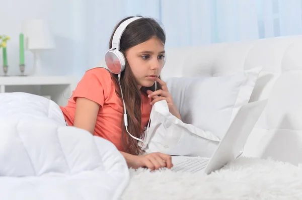 漂亮的小女孩 头戴耳机 软绵绵的米色床铺着笔记本电脑 — 图库照片