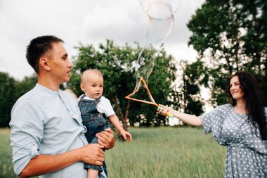 Mutlu genç anne ve baba ve oğul yaz günü çayırda sabun köpüğüyle oynarken eğleniyorlar.