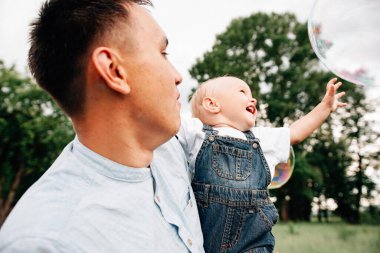 Baba oğlunu elinde tutarken yaz günü çayırda sabun köpüğüyle oynarken. 