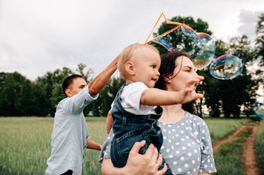 Mutlu genç anne ve baba ve oğul yaz günü çayırda sabun köpüğüyle oynarken eğleniyorlar.