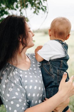 Mutlu, genç bir anne oğlunu elinde tutuyor ve yaz günü çayırda oynarken eğleniyor. 