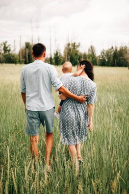 Mutlu, genç ebeveynlerin ve oğullarının yaz günü çayırda durarak kucaklaşmalarının arka görüntüsü. 