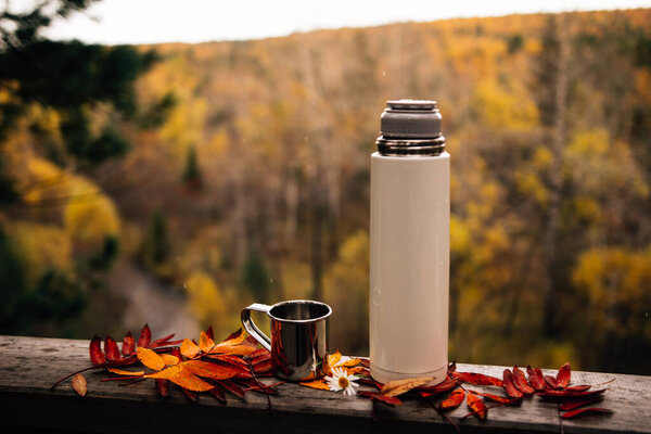 cup with thermos bottle on wooden fence with autumn fallen leaves 