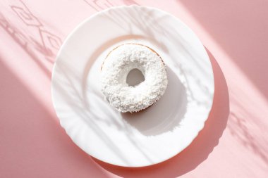 Çörek beyaz sır ile kaplı ve Hindistan cevizi gevreği doğal ışık altında pembe bir arka plan üzerinde beyaz bir tabakta serpilir 