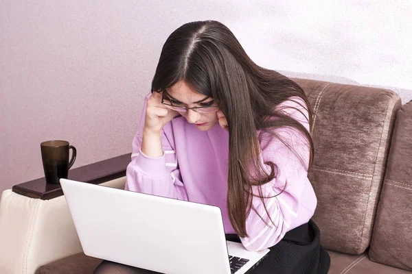 Женщина в очках сидит на коричневом диване и смотрит на ноутбук — стоковое фото