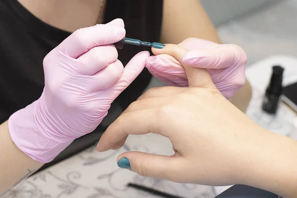 In the nail salon, a manicurist in gloves covers client\'s nails with a green nail Polish with a brush