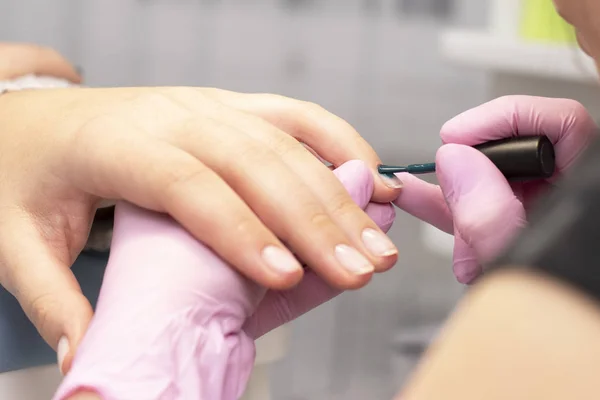 In the nail salon, a manicurist in gloves covers client's nails with a green nail Polish with a brush