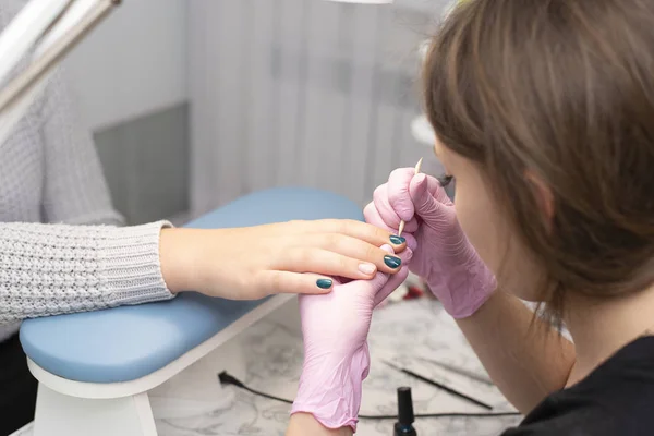 In the nail salon, a manicurist in gloves covers client's nails with a green nail Polish with a brush
