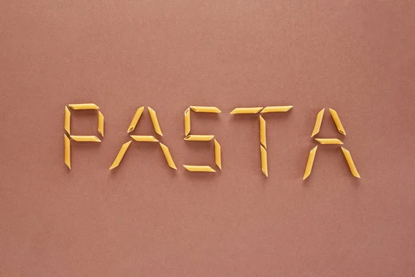 Aufschrift Pasta aus braunen Nudeln in Form von Röhrchen auf braunem Hintergrund — Stockfoto