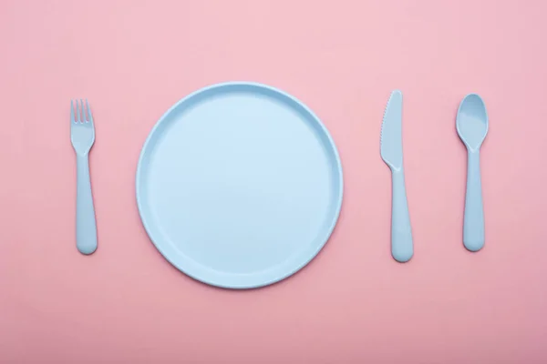 Blue plastic tableware : plate, fork, knife and spoon on pink background.
