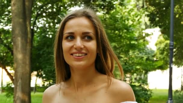 年轻的微笑长发女子模特站在阳光明媚的城市公园微笑 — 图库视频影像