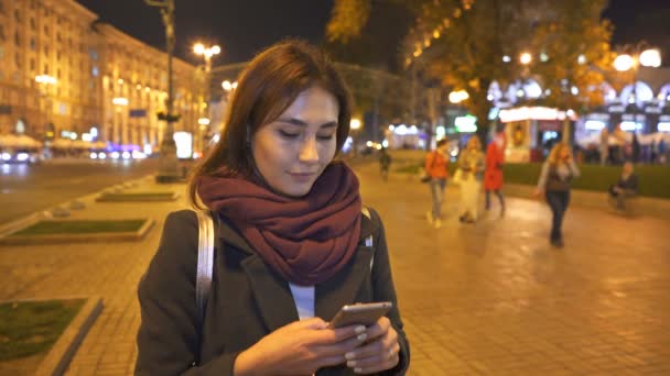 年轻的现代女孩与半亚洲面孔打字智能手机在夜间城市街道 — 图库视频影像