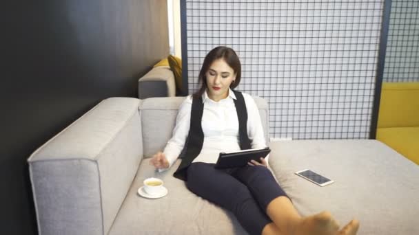 オフィス アジアの若いビジネス ウーマン タブレットとコーヒーが付いているソファーの上に座る 安定したショット — ストック動画