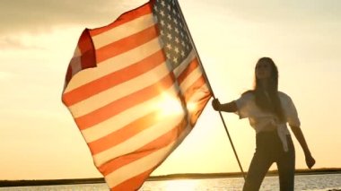 Uzun saçlı kız vatansever genç gökyüzü güneş ile karşı Amerikan bayrağı ile stand. Ağır çekim