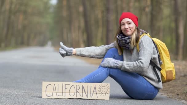 ヒッチハイクの女の子は ポスターカリフォルニアと木の空の道に座っています 指を上向きに — ストック動画