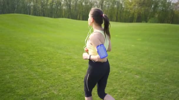 4K緑のフィールドで走る幸せな大人の女性ランナー イヤホンで音楽を聴く — ストック動画