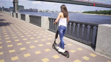 4 k. Kız yavaş yavaş nehir yakınındaki şehir bölgesinde elektrikli scooter sürme. Sabit çekim, geri görünüm