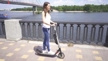 Yavaş çekim. Nehir yakınındaki şehir bölgesinde elektrikli scooter sürme Pretty kız . Sabit atış
