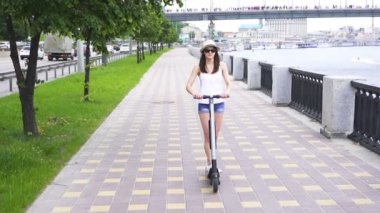 Yavaş çekim. Nehir yakınındaki şehir bölgesinde şapkalı kız elektrikli scooter sürüyor. Sabit atış