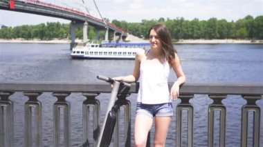 Yavaş çekim. Mutlu genç esmer kadın şehir bölgesinde elektrikli scooter yakınında durmak. Sabit atış
