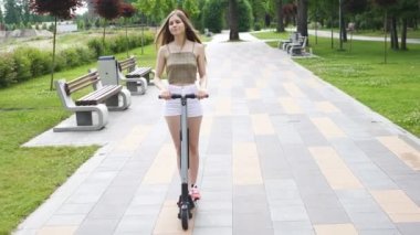 4 k. Genç uzun saçlı kız yavaş yavaş şehir parkı alanında elektrikli scooter sürme. Sabit atış, ileri görüş