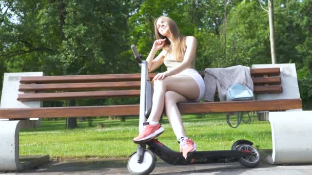 Šťastná usměvavá mladá brunetka sedí na lavičce u elektrického skútru v oblasti městského parku. Stálý portrét