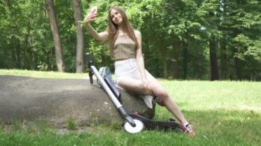 Ağır çekim. Şehir parkında elektrikli scooter kullanan genç seksi kız. Arka plan