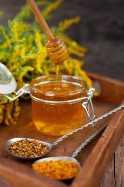 Cam kavanozda tahta kaşıkla bal. Sarı çiçekli, ahşap bir masada altın sarısı bir çubuk. Sağlıklı tatlı yiyecekler.