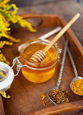 Cam kavanozda tahta kaşıkla bal. Sarı çiçekli, ahşap bir masada altın sarısı bir çubuk. Sağlıklı tatlı yiyecekler.
