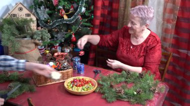 Büyükannem Noel ağacı çelengi yapar. Yaşlı bir kadın. Noel ağacı topları ve oyuncaklarla süslenmiş. Kırmızı elbise. Kış aktiviteleri. Aralık 'ta memlekette.