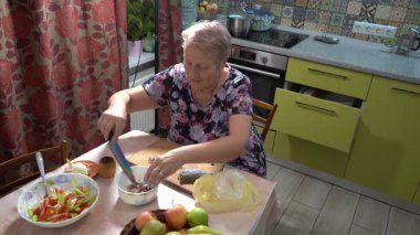 Yaşlı bir kadın mutfakta balık yemi pişiriyor. Tahtayı kesiyor. Ringa. Sebze salatası.