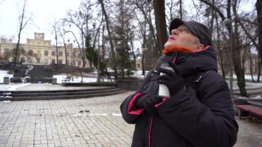 Yaşlı bir kadın caddedeki bir fincandan kahve içer. Parkta kar var. Bir büyükanne kışın doğada yürür. Açık hava..