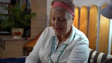 Yaşlı, gülümseyen bir kadın mücevherlerini deniyor. Gri saçlı bir büyükanne zincirine kolye takar. Evde yatakta otur. Boynunda boncuklar var. Mücevherler